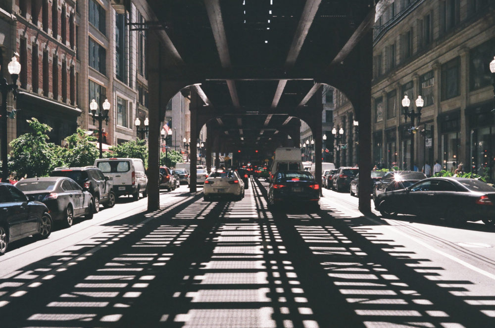 cars drive through the Chicago loop, in the shadows of overhead transit rail lines