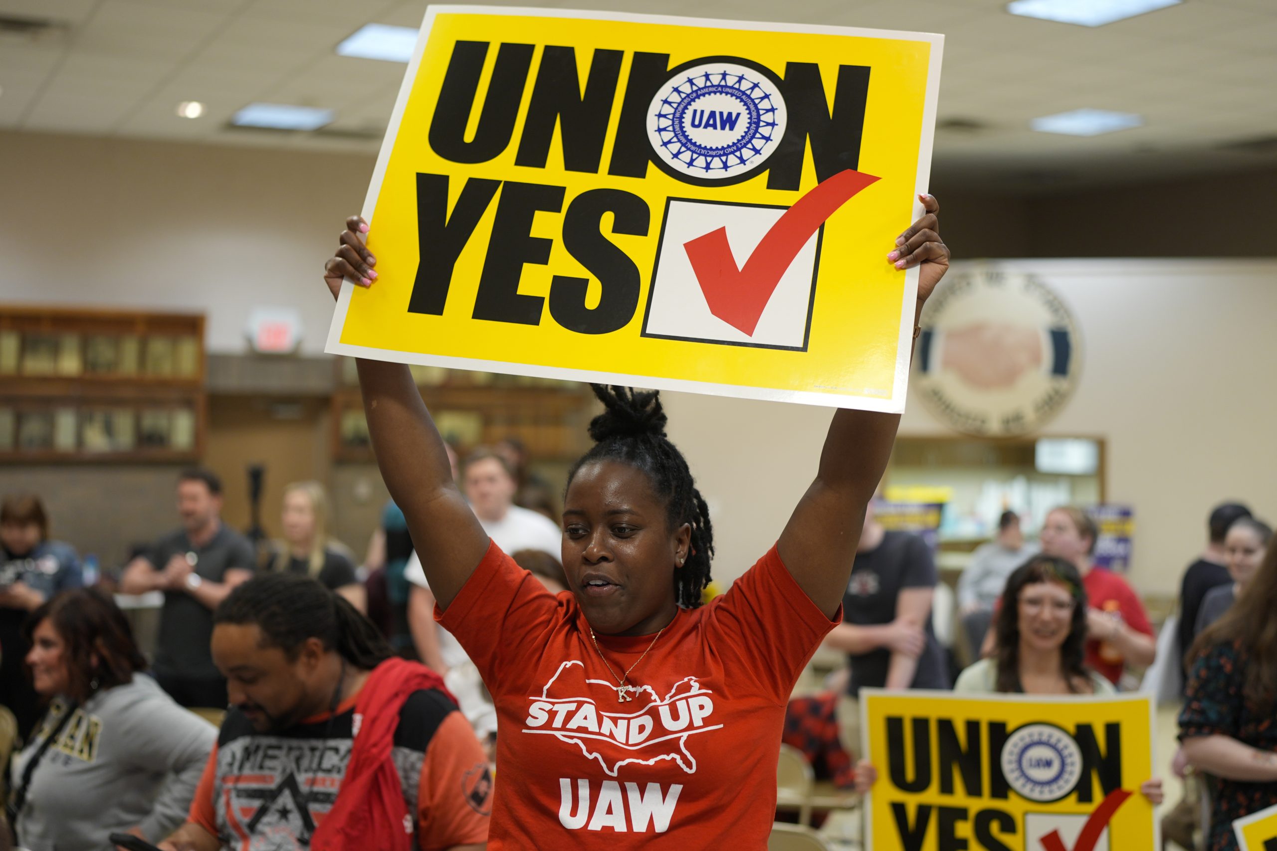 A woman in a red UAW t-shirt holds a yellow sign reading "Union Yes" over her head