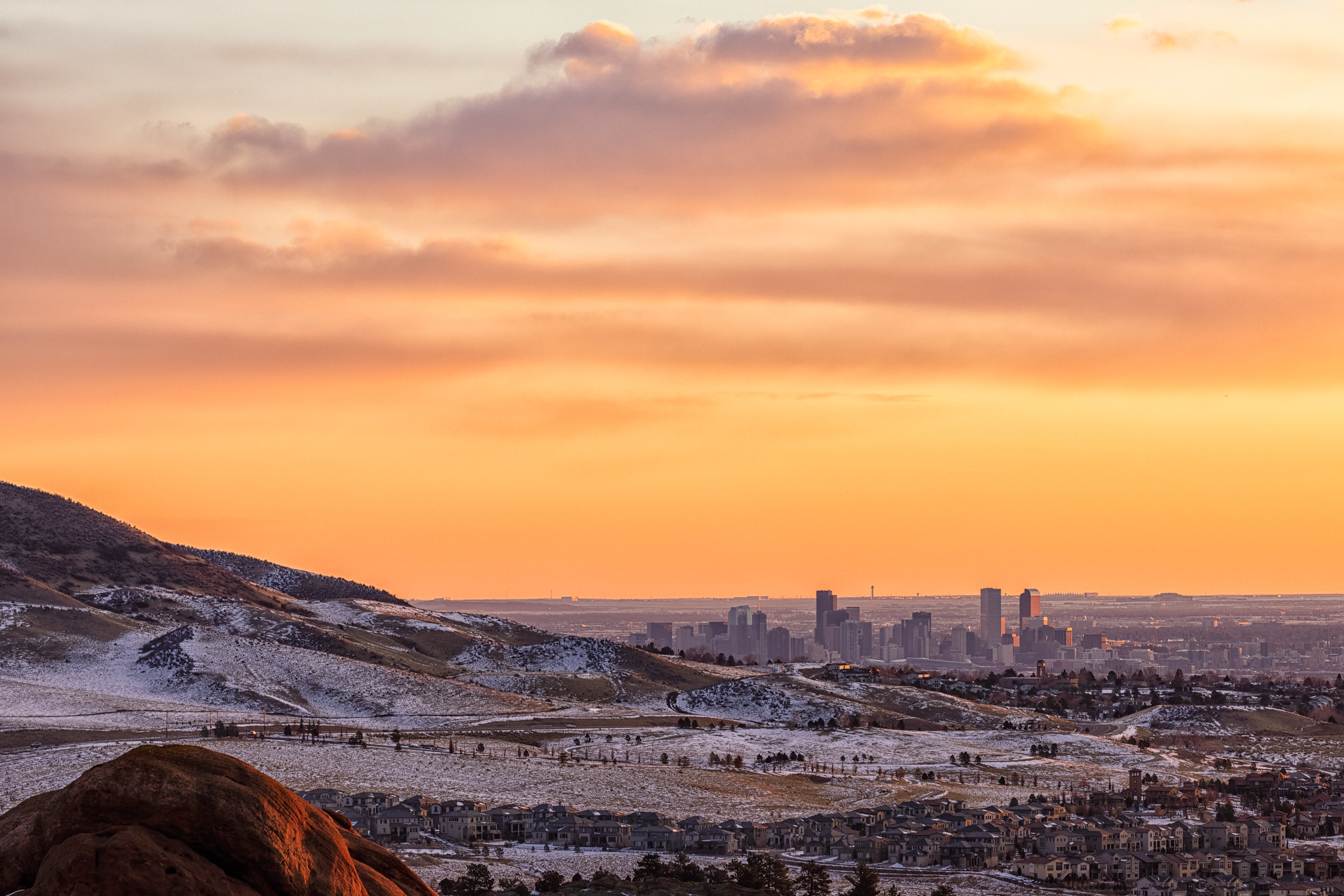 A distant view of Denver, Colorado showing suburban homes encroaching on the Rocky Mountain foothills with a yellow sunset in the background.