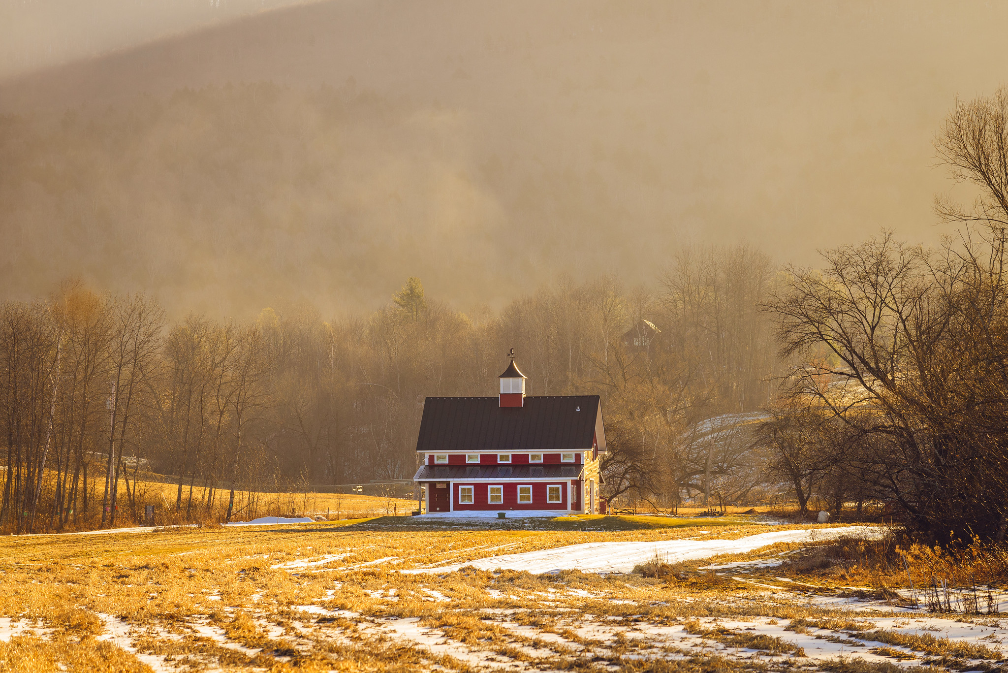 A large red barn sits in a golden field streaked with just a bit of snow
